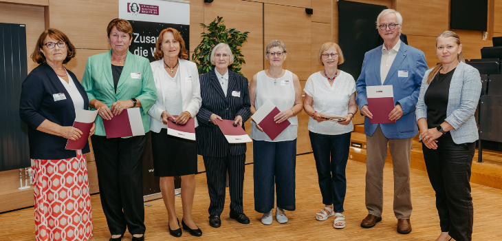 Acht Personen stehen nebeneinander in einem Hörsaal und halten Urkunden in den Händen. Sie tragen festliche Kleidung und lächeln in die Kamera. Im Hintergrund ist ein Banner der Universität zu sehen ©Jana Dünnhaupt, Uni Magdeburg