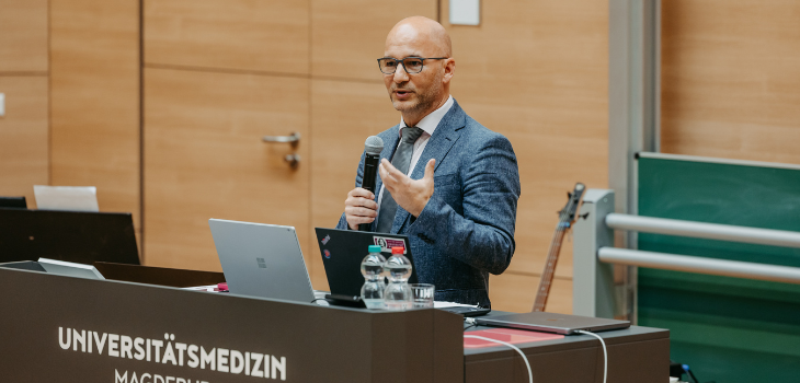 Ein Mann mittleren Alters steht an einem Rednerpult mit der Aufschrift „Universitätsmedizin Magdeburg“. Er trägt einen grauen Anzug, eine Krawatte und eine Brille. Mit einem Mikrofon in der Hand spricht er zu den Gästen. Auf dem Pult stehen ein Laptop und zwei Wassergläser ©Jana Dünnhaupt, Uni Magdeburg